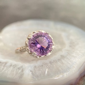 Może przedstawiać: Srebrny pierścionek z dużym, okrągłym, fioletowym ametystem osadzonym w ozdobnej, srebrnej oprawie.