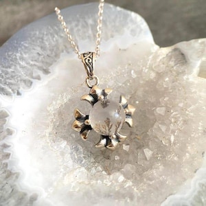 May include: A silver pendant necklace featuring a floral design with a clear, round stone at the center. The pendant is suspended from a delicate silver chain. The necklace is displayed on a white and gray stone surface.