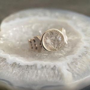 May include: A silver ring with a round, clear stone featuring a delicate, fern-like pattern. The ring has a unique design with decorative elements on the band. The ring is displayed on a white and gray surface.