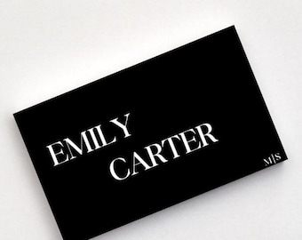 May include: A rectangular black business card featuring the white printed text EMILY CARTER in a serif font. The letters M|S are printed in a smaller white serif font in the bottom right corner of the card, which is positioned at an angle.