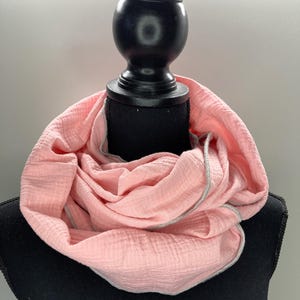 Könnte beinhalten: Ein rosa Baumwollschal mit weißem Rand. Der Schal ist gefaltet und auf einer schwarzen Schaufensterpuppe ausgestellt.