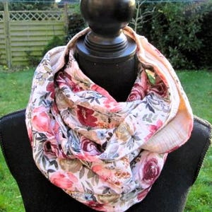 Könnte beinhalten: Ein floraler Loop-Schal auf einer schwarzen Schaufensterpuppe. Der Schal hat ein Muster aus rosa, roten und weißen Blumen mit grünen Blättern. Der Hintergrund ist eine verschwommene Ansicht eines grünen Rasens und eines Holzzauns.