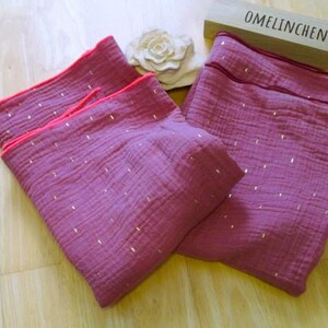Könnte beinhalten: Zwei rosa Musselin-Swaddles mit goldenen Glitzerpunkten und rosa Besatz. Die Swaddles sind gefaltet und auf einer Holzoberfläche ausgelegt. Der Text "OMELINCHEN" ist auf einem Holzblock im Hintergrund sichtbar.