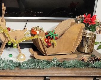 May include: A wooden Christmas sleigh and reindeer decoration, adorned with gold tinsel, ornaments, and wrapped gifts. The sleigh is filled with presents and sits on a wooden surface, with a garland of green tinsel.