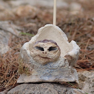 Puede incluir: Una escultura de cerámica blanca de una cara con ojos grandes y una boca pequeña. La escultura está sentada en un pequeño cuenco de cerámica blanca.
