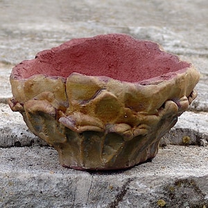 May include: A handcrafted, textured ceramic bowl with a rough, organic shape. The interior is a deep red, contrasting with the earthy tones of the exterior, which features shades of tan, brown, and green. The bowl sits on a weathered stone surface.