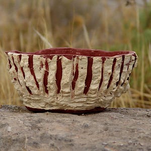 Puede incluir: Un cuenco de cerámica hecho a mano con un exterior texturizado blanquecino y rayas rojas verticales. El interior es de color rojo liso. El cuenco está sobre una superficie de piedra gris, con un fondo natural borroso.