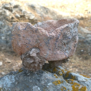 Puede incluir: Una olla de barro rojizo y desgastado con una superficie rugosa y desigual. La olla está sentada sobre una superficie rocosa con algo de musgo verde.