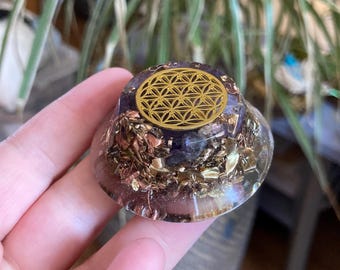 Può includere: Una piramide di orgone in resina trasparente con scaglie di metallo color oro e una base di pietra viola. La parte superiore presenta una sovrapposizione di metallo color oro con un design geometrico. La piramide è tenuta in mano.
