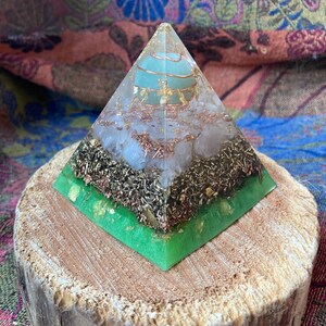 Peut inclure: Une pyramide en cristal verte, blanche et dorée avec une sphère en cristal transparent au sommet. La pyramide est posée sur une base en bois.