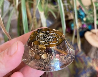 Può includere: Una piramide di orgone trasparente a forma di cupola con un simbolo di Geometria Sacra color argento sulla parte superiore. La piramide contiene pietre occhio di tigre e altri elementi metallici. La base è tenuta in mano, con piante verdi sullo sfondo.