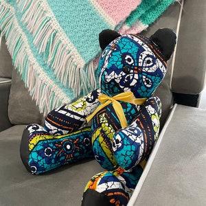 May include: A blue and white patterned teddy bear with black ears and a yellow ribbon around its neck. The bear is sitting on a gray armchair.