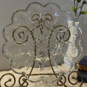 May include: A clear glass serving dish with a scalloped edge and a starburst pattern in the center. The dish is on a gold metal stand.