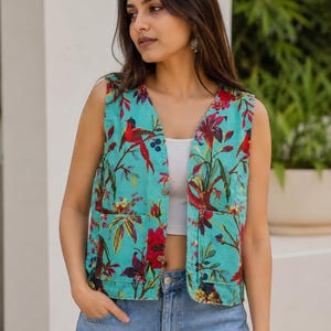 May include: A sleeveless vest in a vibrant turquoise color featuring a colorful botanical print with red birds and floral patterns. The vest has a V-neckline, a front button closure with small brown buttons, and two square patch pockets on the front.