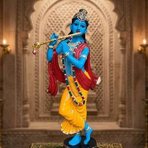 May include: A blue-skinned deity statue standing on a black circular base, wearing a yellow dhoti and red shawl. The figure holds a gold flute to their lips, adorned with a silver crown, necklaces, and armbands. The text Ayzleen Treasure is visible.