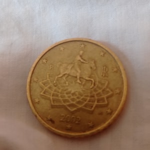 Peut inclure: Une pièce de monnaie dorée représentant une personne à cheval, entourée d'étoiles et d'un motif floral. L'année 2002 est visible. La pièce porte la lettre "R" des deux côtés.