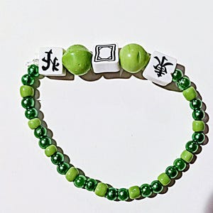 May include: A green beaded bracelet with white square charms bearing black characters and green round beads. The bracelet features light and dark green beads, creating a vibrant accessory.