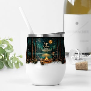 May include: A white stainless steel tumbler with a lid and a white straw, featuring a printed graphic of a forest scene at night with a campfire, two chairs, a full moon, and the text Life is better by the campfire in gold lettering.