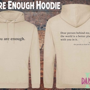 May include: Beige hoodie with the text "You are enough" on the front and a message on the back. The hoodie features a drawstring hood and a front pocket. The back of the hoodie says "Dear person behind me, the world is a better place with you in it."