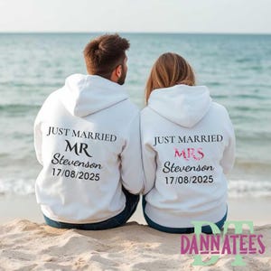 May include: Two white hoodies with the text "JUST MARRIED" and the names "MR Stevenson" and "MRS Stevenson" printed on the back. The date "17/08/2025" is printed below the names. The couple is sitting on a sandy beach.