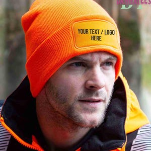 Puede incluir: Gorro naranja brillante con un parche rectangular que dice "YOUR TEXT / LOGO HERE". El gorro es de punto. La persona de la imagen lleva una chaqueta negra y naranja.