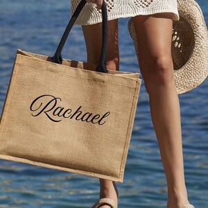 Op de afbeelding: Een bruine jute draagtas met zwarte handvatten. De naam "Rachael" staat in sierlijke letters op de voorkant. Een strohoed wordt in de buurt gehouden. De achtergrond is een wazig uitzicht op de zee.