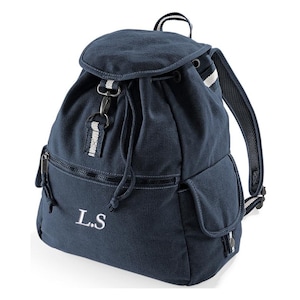 Può includere: Zaino in tela blu navy con chiusura a coulisse e patta. Presenta una tasca frontale con cerniera, una tasca laterale e spallacci regolabili. Lo zaino ha dettagli a righe bianche e grigie e le iniziali "L.S" ricamate sul davanti.