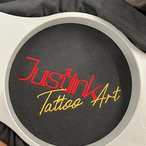以下が含まれることがあります： 黒いTシャツのアップで、赤い糸と金色の糸で「Just Ink Tattoo Art」と刺繍されています。円形の枠の中に刺繍されています。