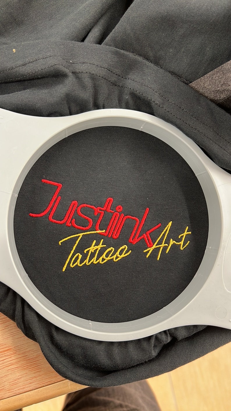 以下が含まれることがあります： 黒いTシャツのアップで、赤い糸と金色の糸で「Just Ink Tattoo Art」と刺繍されています。円形の枠の中に刺繍されています。