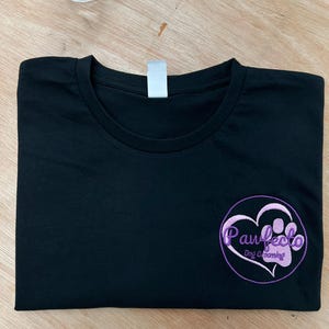 以下が含まれることがあります： パウフェクトドッググルーミングと刺繍された紫色のロゴがついた黒いTシャツ。ロゴは、足跡とハートが描かれた円の中にあります。