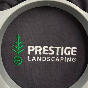 以下が含まれることがあります： グレーのTシャツのクローズアップ。緑色の木のロゴと「PRESTIGE LANDSCAPING」という文字が白で刺繍されています。