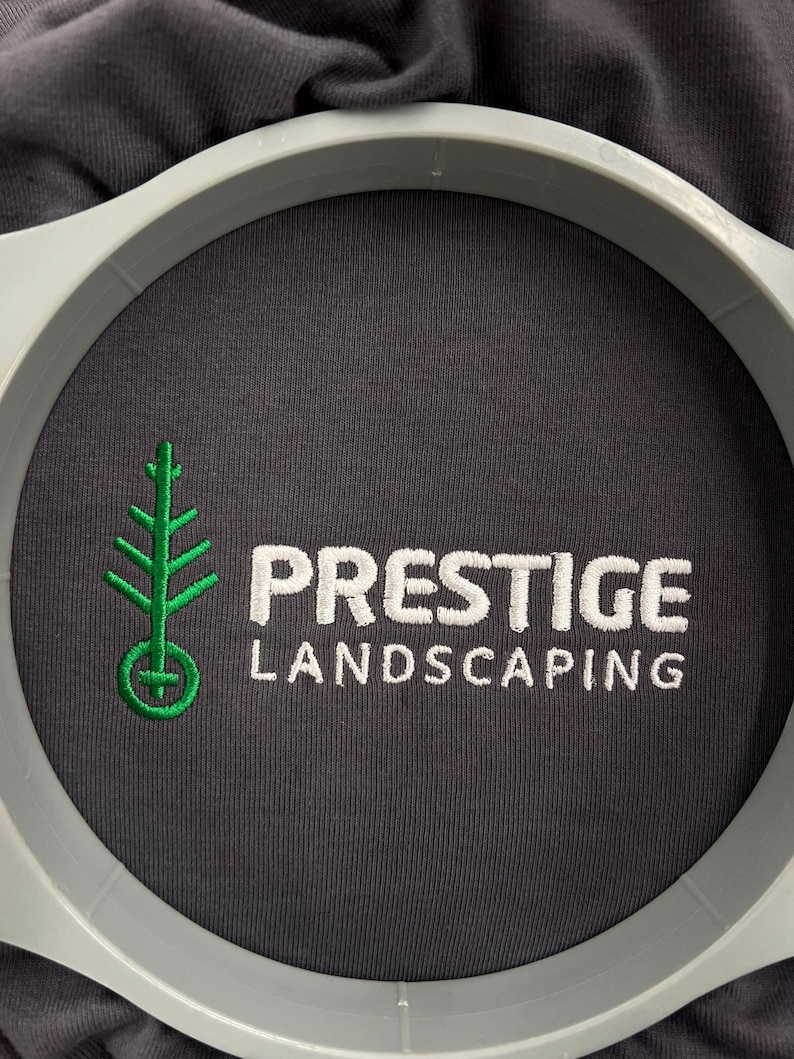 以下が含まれることがあります： グレーのTシャツのクローズアップ。緑色の木のロゴと「PRESTIGE LANDSCAPING」という文字が白で刺繍されています。