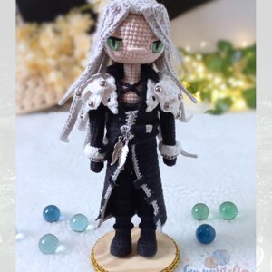 May include: Crocheted doll of a character with long silver hair, green eyes, and a black coat with white accents. The doll is standing on a wooden base with gold trim, surrounded by glass beads. The doll's design includes a feather pendant.