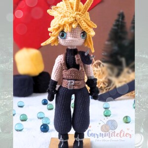 May include: A handmade crochet doll of a character with spiky yellow hair, blue eyes, and black gloves. The doll wears a black top, navy blue pants, and a tan vest with brown straps. The doll is standing on a wooden base. The doll is a Final Fantasy character.