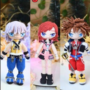 Puede incluir: Tres muñecas de ganchillo hechas a mano inspiradas en los personajes de Kingdom Hearts. Cada muñeca tiene atuendos y peinados únicos, con colores vibrantes como el azul, el rojo y el amarillo. Las muñecas se exhiben sobre una superficie blanca.