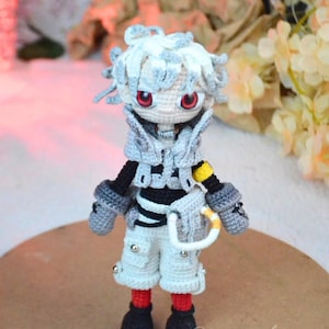 May include: A handmade crochet doll with white hair and red eyes. The doll wears a gray vest, black and white striped shirt, white shorts, and black boots. A yellow and white accessory hangs from the doll's waist. The doll is a unique, handcrafted item.