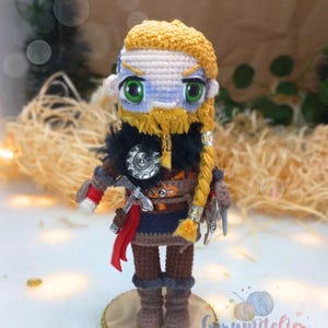 May include: A crocheted figurine of a warrior with a blonde braid, wearing a blue and brown tunic, holding a silver axe and a shield. The figurine is standing on a wooden base with a gold trim.