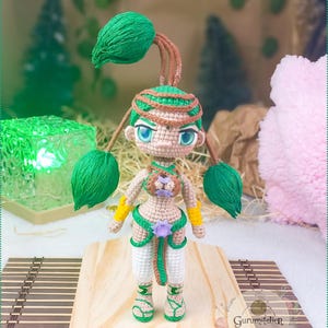 Può includere: Bambola all'uncinetto fatta a mano con capelli e dettagli verdi, indossa un completo beige e verde con bracciali gialli e pantaloni bianchi. La bambola è in piedi su una superficie di legno. La bambola ha un fiore viola in vita.