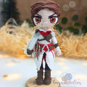 May include: Crocheted amigurumi doll of a character wearing a white hooded cloak, red sash, and brown boots. The doll has brown hair, red eyes, and a serious expression. The doll is standing on a wooden base with a gold trim.