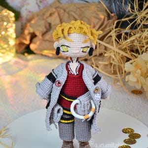 May include: A handmade crochet doll with yellow hair, wearing a gray coat over a red sweater and gray pants. The doll has yellow eyes and black earrings. A white and orange ring hangs from the doll's waist. Gold coins are scattered around the doll.