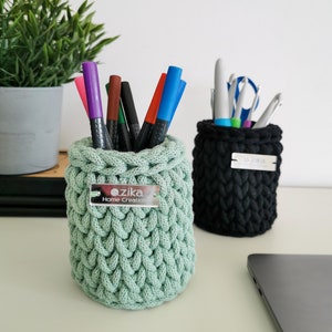 Könnte beinhalten: Zwei handgefertigte Stifthalter aus Häkelarbeit, einer hellgrün und einer schwarz, stehen auf einem Schreibtisch. Der hellgrüne Halter enthält bunte Marker, während der schwarze Schere und Stifte enthält. Jeder Halter hat ein Metallschild mit der Aufschrift "zika Home Creations".