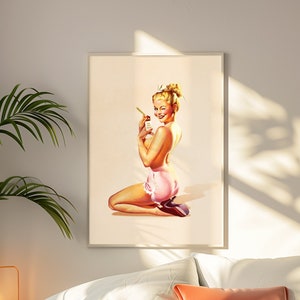 May include: A framed wall art print featuring a blonde pin-up figure kneeling while wearing pink ruffled shorts and dark heels. The figure holds a small bottle and looks over their shoulder with a wink against a light cream background.