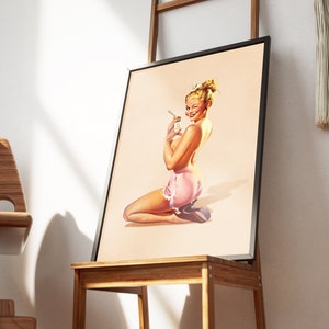 May include: A framed art print featuring a blonde person with an updo hairstyle, kneeling while wearing a light pink skirt with white lace trim. The person is winking and holding a small white rectangular object with text in their hand.