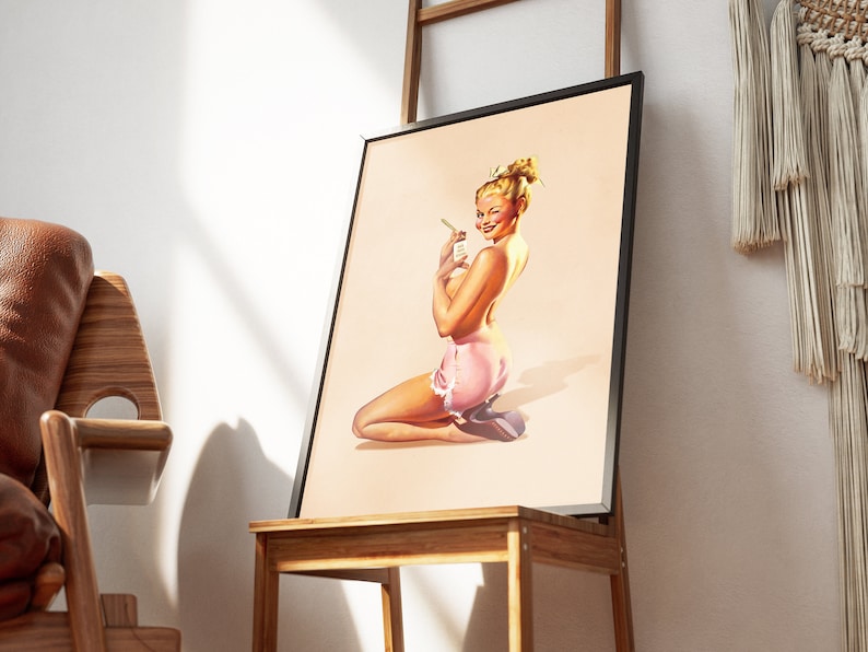 May include: A framed art print featuring a blonde person with an updo hairstyle, kneeling while wearing a light pink skirt with white lace trim. The person is winking and holding a small white rectangular object with text in their hand.