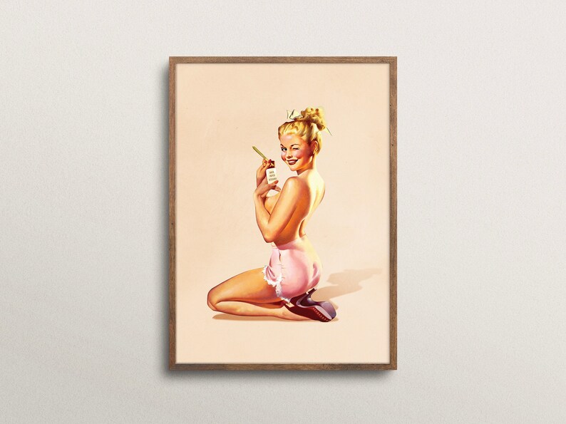 May include: A framed art print featuring a blonde person with an updo hairstyle, kneeling while wearing pink shorts with white lace trim. The person is winking and holding a small rectangular object with text in their hand.