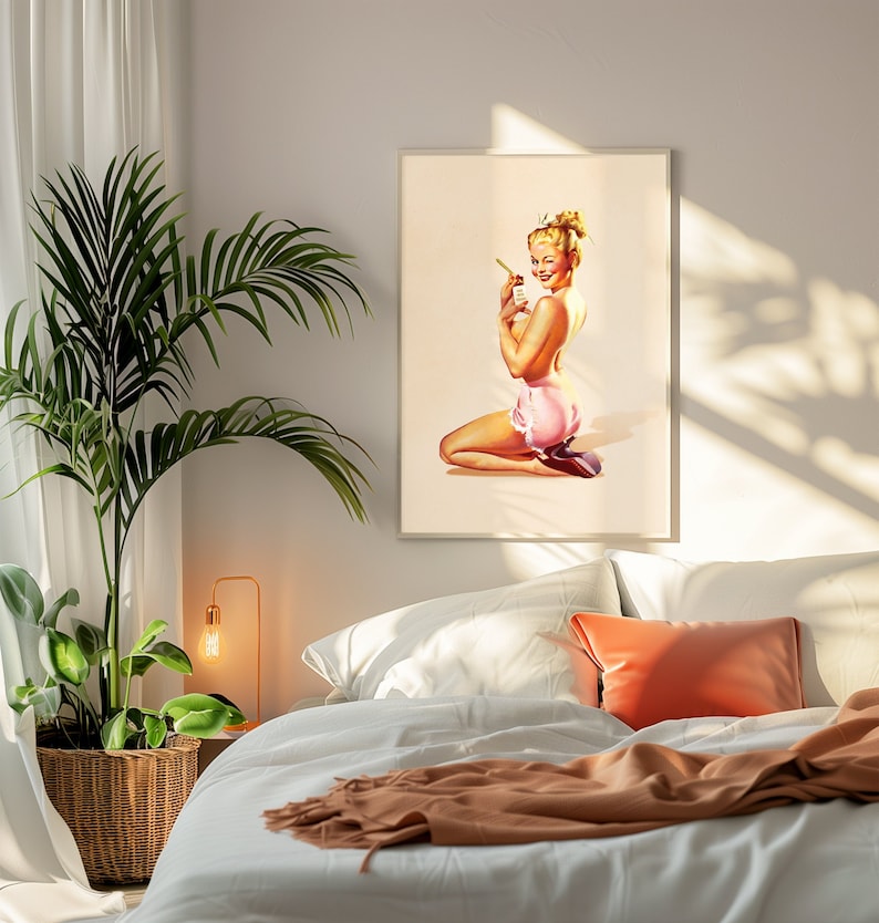 May include: A framed wall art print featuring a blonde pin-up figure kneeling while wearing pink ruffled shorts and dark heels. The figure holds a small bottle and looks over their shoulder with a wink against a light cream background.