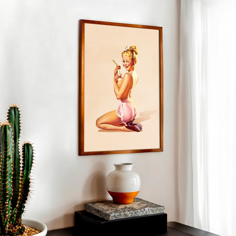 May include: A framed wall art print featuring a vintage-style illustration of a blonde person with an updo hairstyle, kneeling while wearing pink ruffled shorts and holding a small bottle. The artwork is set against a light beige background.