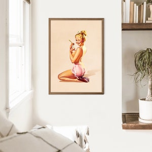 May include: A framed wall art print featuring a vintage-style illustration of a blonde person with an updo hairstyle, kneeling while wearing pink ruffled shorts and holding a small bottle. The artwork has a light beige background and a brown frame.