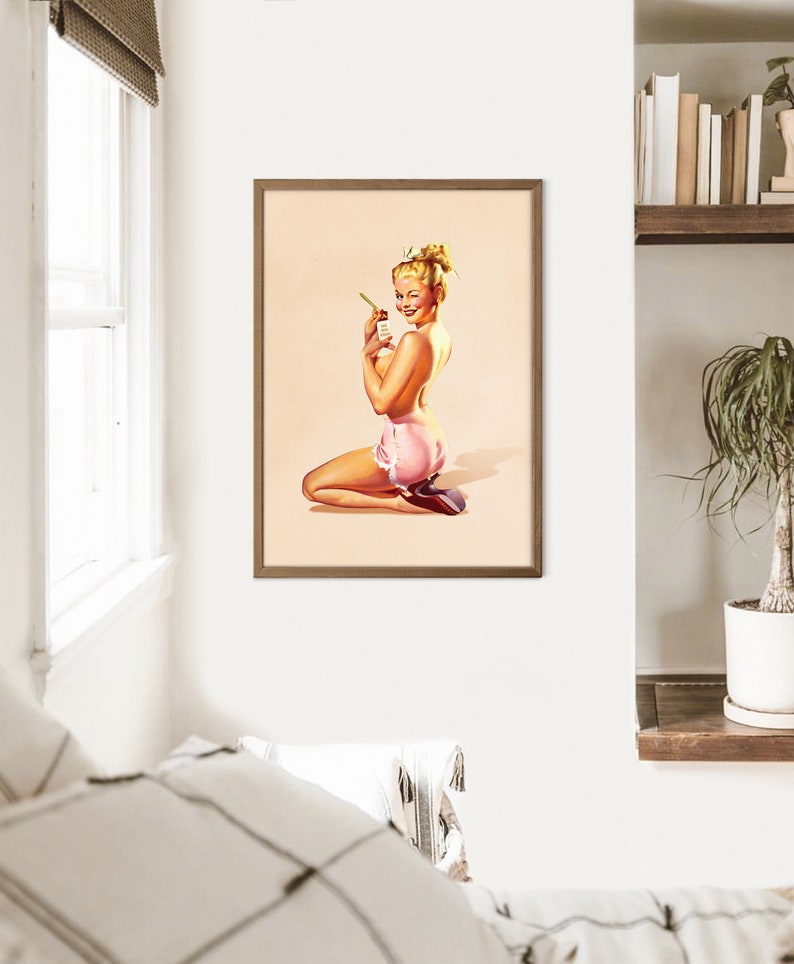 May include: A framed wall art print featuring a vintage-style illustration of a blonde person with an updo hairstyle, kneeling while wearing pink ruffled shorts and holding a small bottle. The artwork has a light beige background and a brown frame.