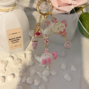 Puede incluir: Un llavero dorado con múltiples dijes. Los dijes incluyen una tetera rosa, una rosquilla rosa, una bebida rosa y una pequeña tetera blanca. El llavero está decorado con cuentas rosas y un cierre dorado.
